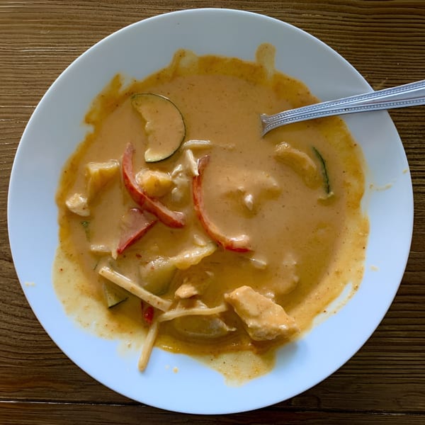 A white plate of Spikey Ty's Asian curry with chicken, vegetables, and a spoon on a wooden table.